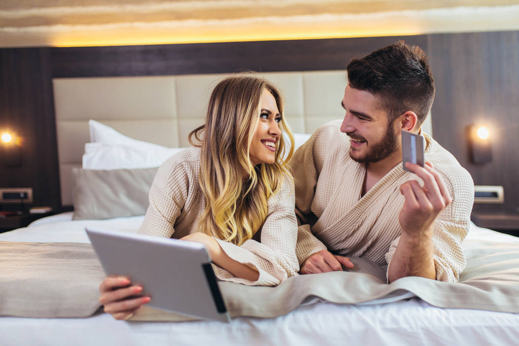 Happy couple using digital tablet and credit card while lying together in bed in hotel room.