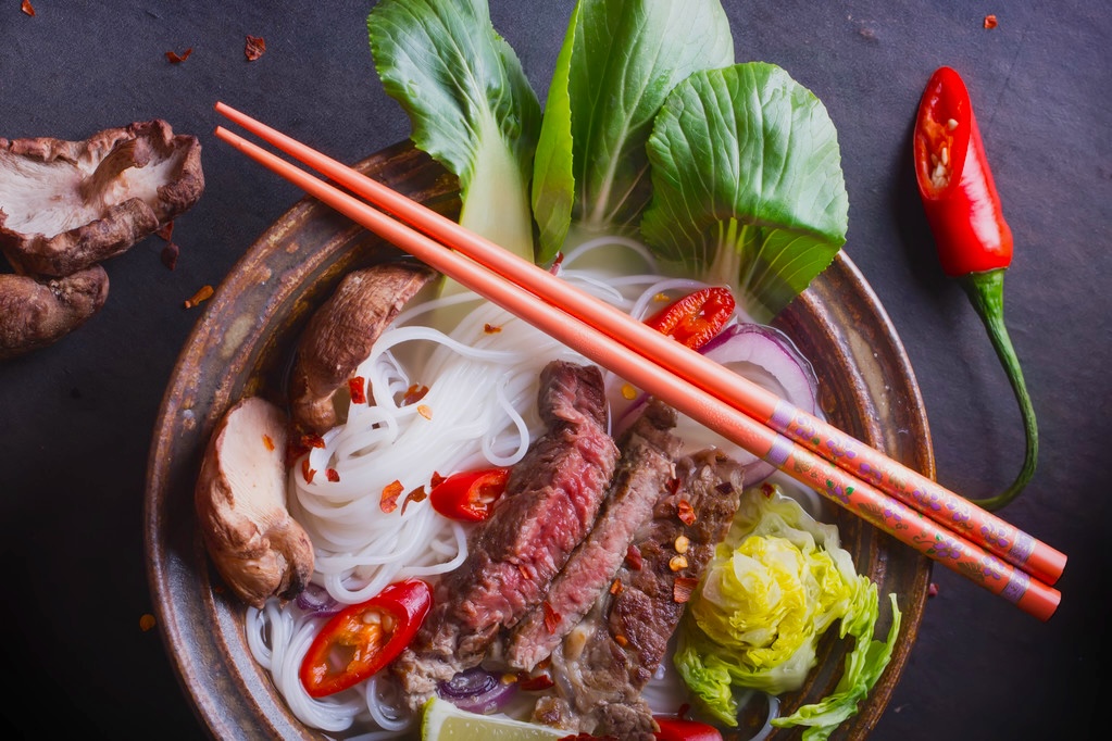 Vietnamese Pho Bo soup served in vintage oriental bowl with chop sticks, seasoning and greens on background